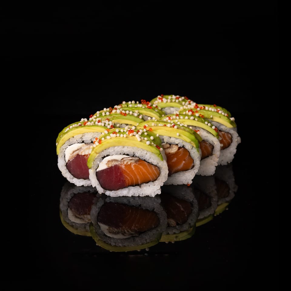 Assorted sushi rolls filled with salmon and tuna, topped with avocado slices and red and white roe on a reflective black surface.