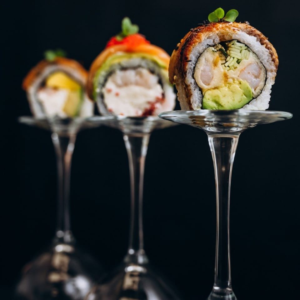 Three sushi rolls with colorful fillings and green garnishes displayed on elegant cocktail glasses against a dark background.