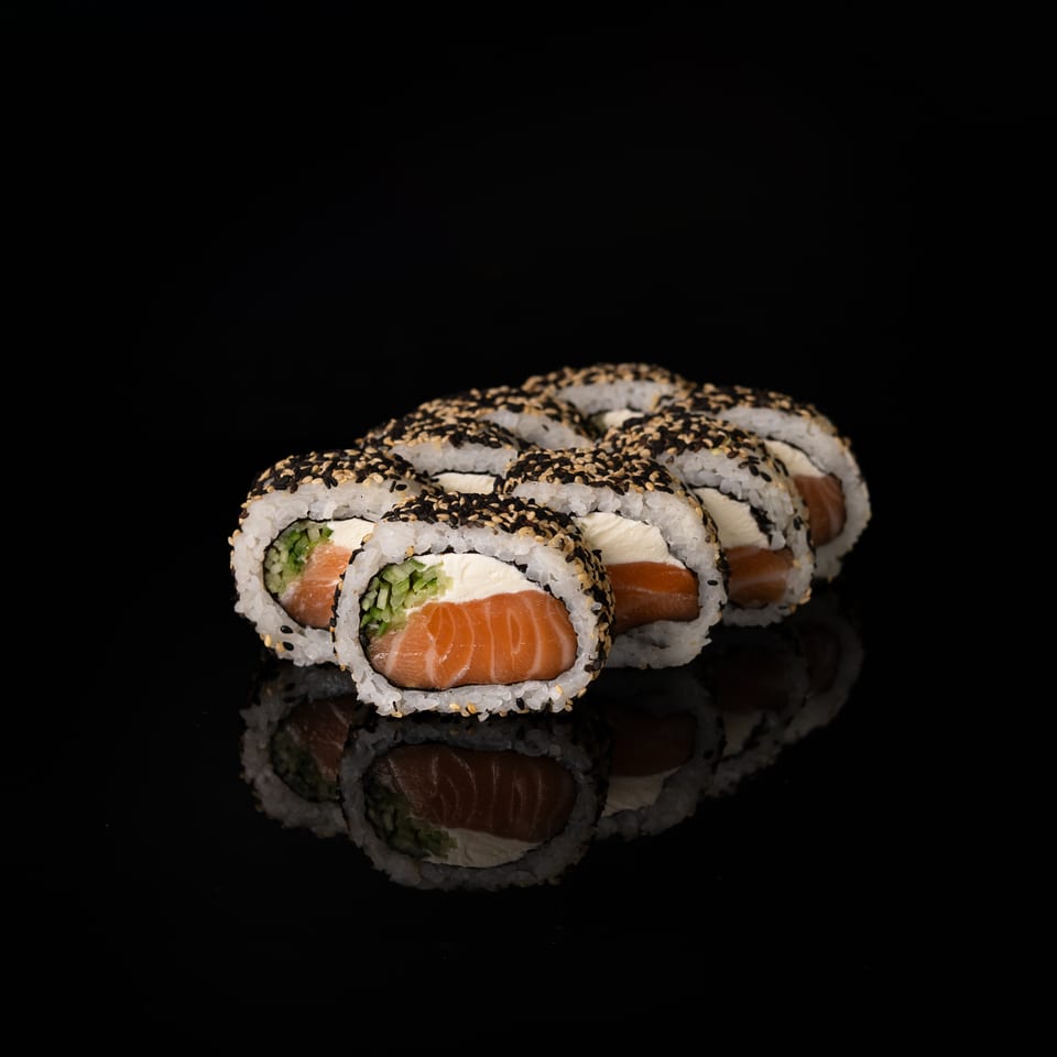 Sushi rolls filled with salmon, cucumber, and cream cheese, coated in black and white sesame seeds on a reflective black surface.