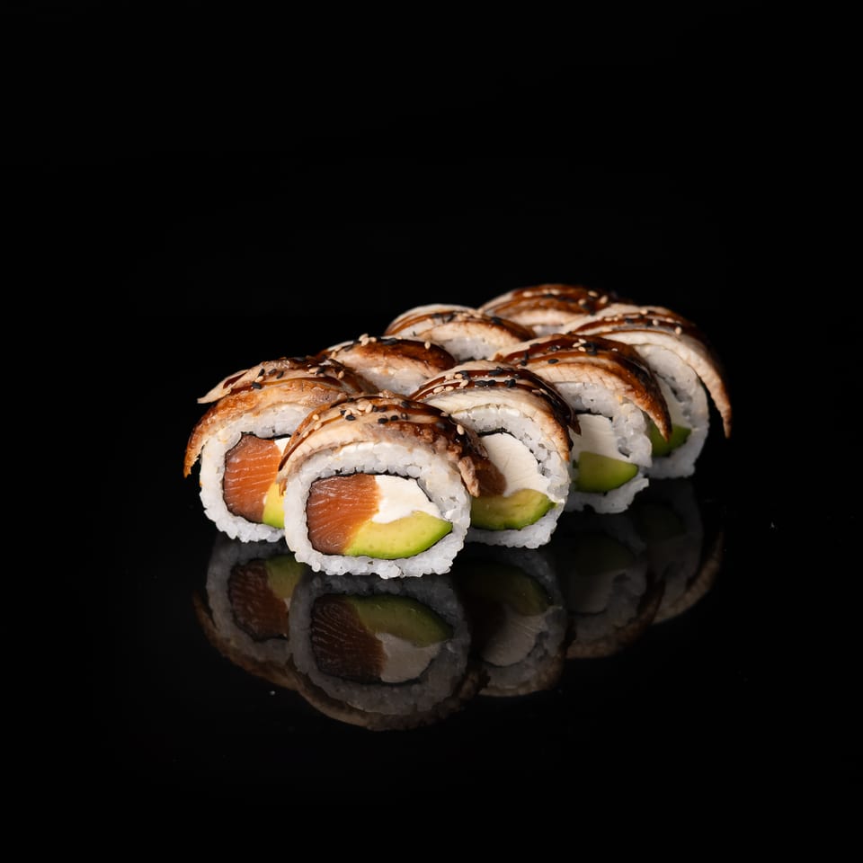 Sushi rolls with salmon, avocado, and cream cheese topped with seared fish and sesame seeds on a reflective black surface.